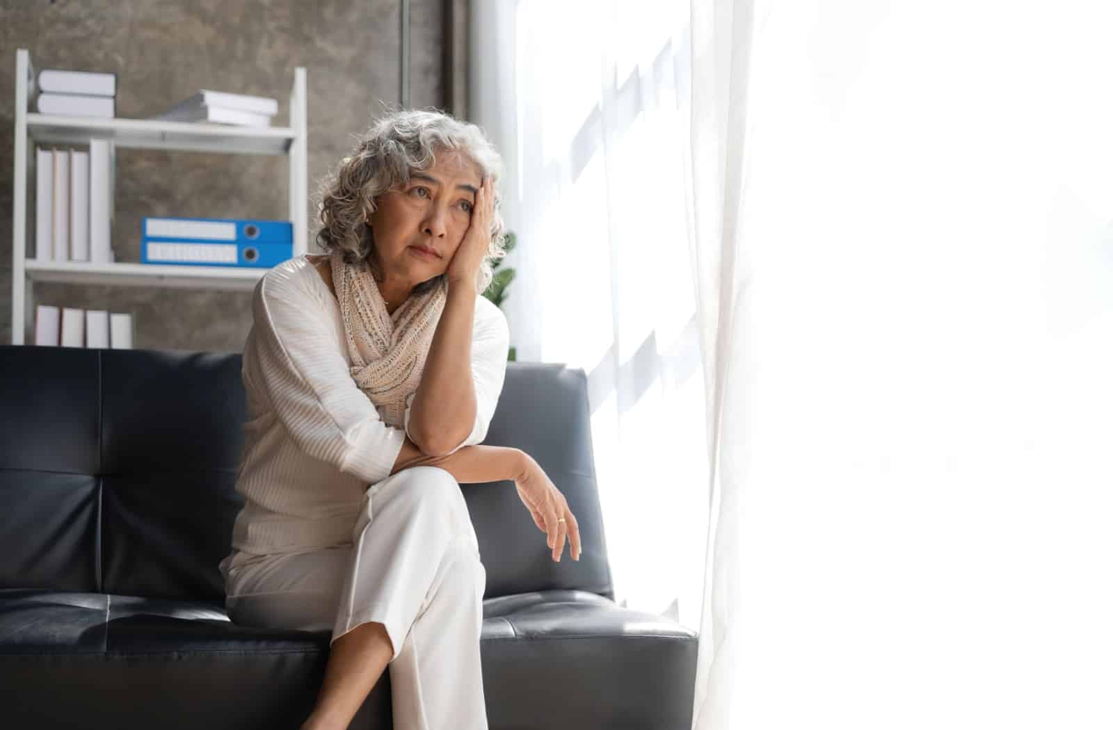 A senior holds a hand to their head as they look worryingly out of an open window while sitting on a black couch