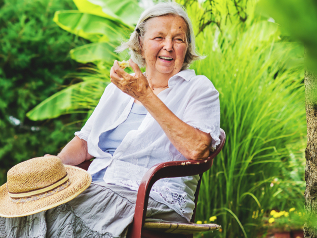 Smiling elderly woman in a white blouse sits on a garden chair, holding a leaf with a straw hat on her lap.